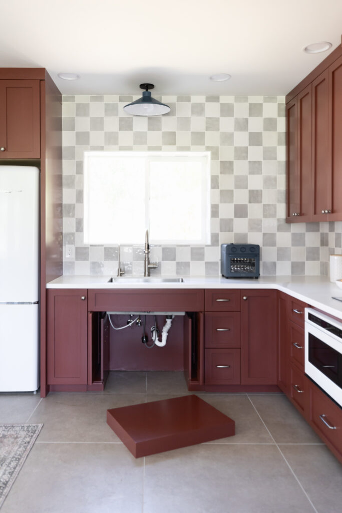 Accessible roll-under kitchen sink with removable base for wheelchair users, featuring deep red cabinetry, white countertops, and a gray checkered tile backsplash.