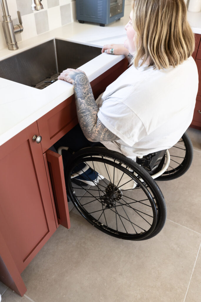 Accessible roll-under kitchen sink with removable base for wheelchair users, featuring deep red cabinetry, white countertops, and a gray checkered tile backsplash. Blonde woman in a wheelchair using a roll under. 