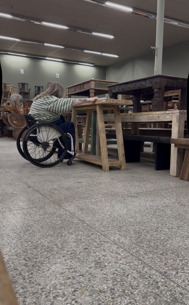 Wheelchair user testing table height and furniture clearance inside a vintage furniture warehouse while evaluating accessibility and transfer space for interior design.