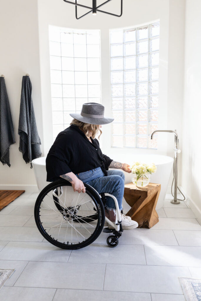 Wheelchair user in a bright, modern accessible bathroom reaching toward a small wood stool with flowers beside a freestanding tub, showcasing barrier free design, wide clearances, and stylish universal design details.