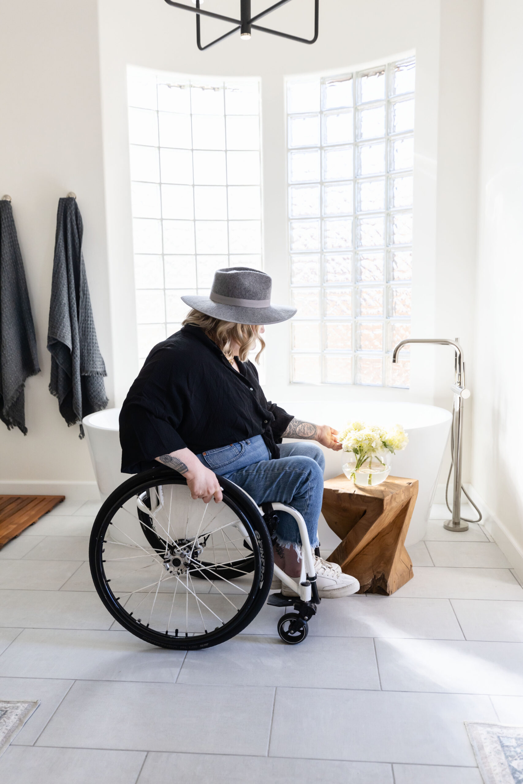 Wheelchair user in a bright, modern accessible bathroom reaching toward a small wood stool with flowers beside a freestanding tub, showcasing barrier free design, wide clearances, and stylish universal design details.
